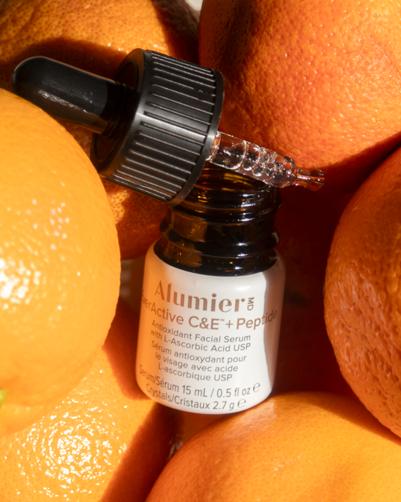 Close-up of a dark dropper bottle labeled Alumier among several bright orange fruits (oranges).