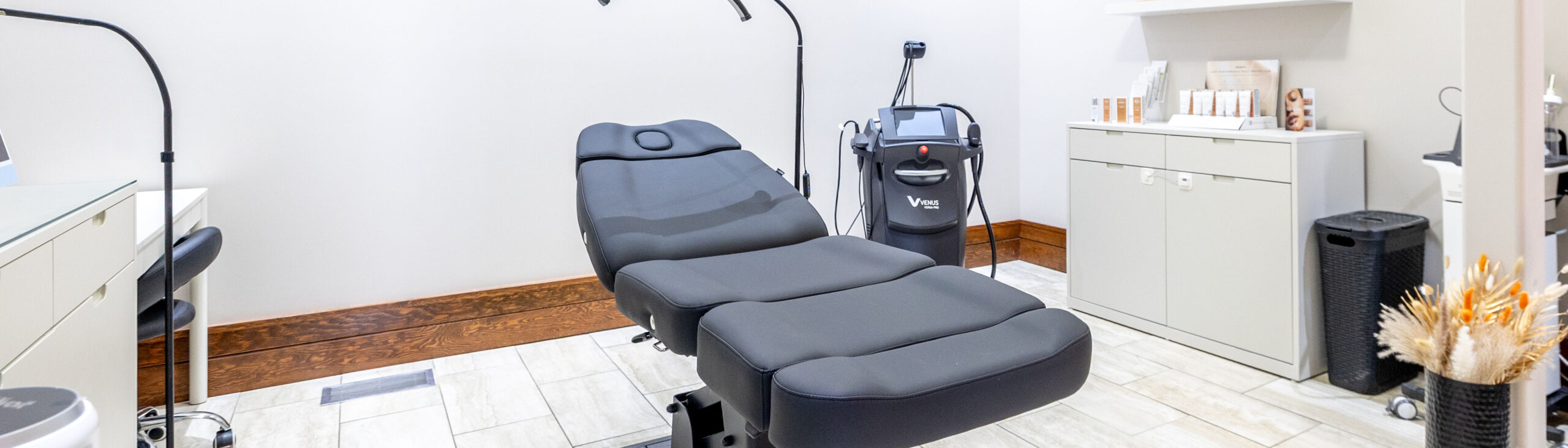Gray padded reclining treatment chair in a bright clinic room with skincare equipment and storage cabinets left and right